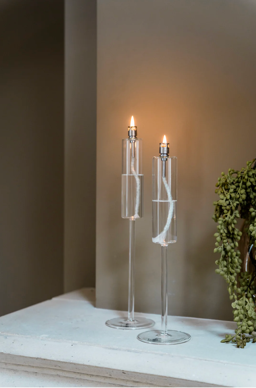 Two lit candles in clear glass holders on a white surface with a neutral background