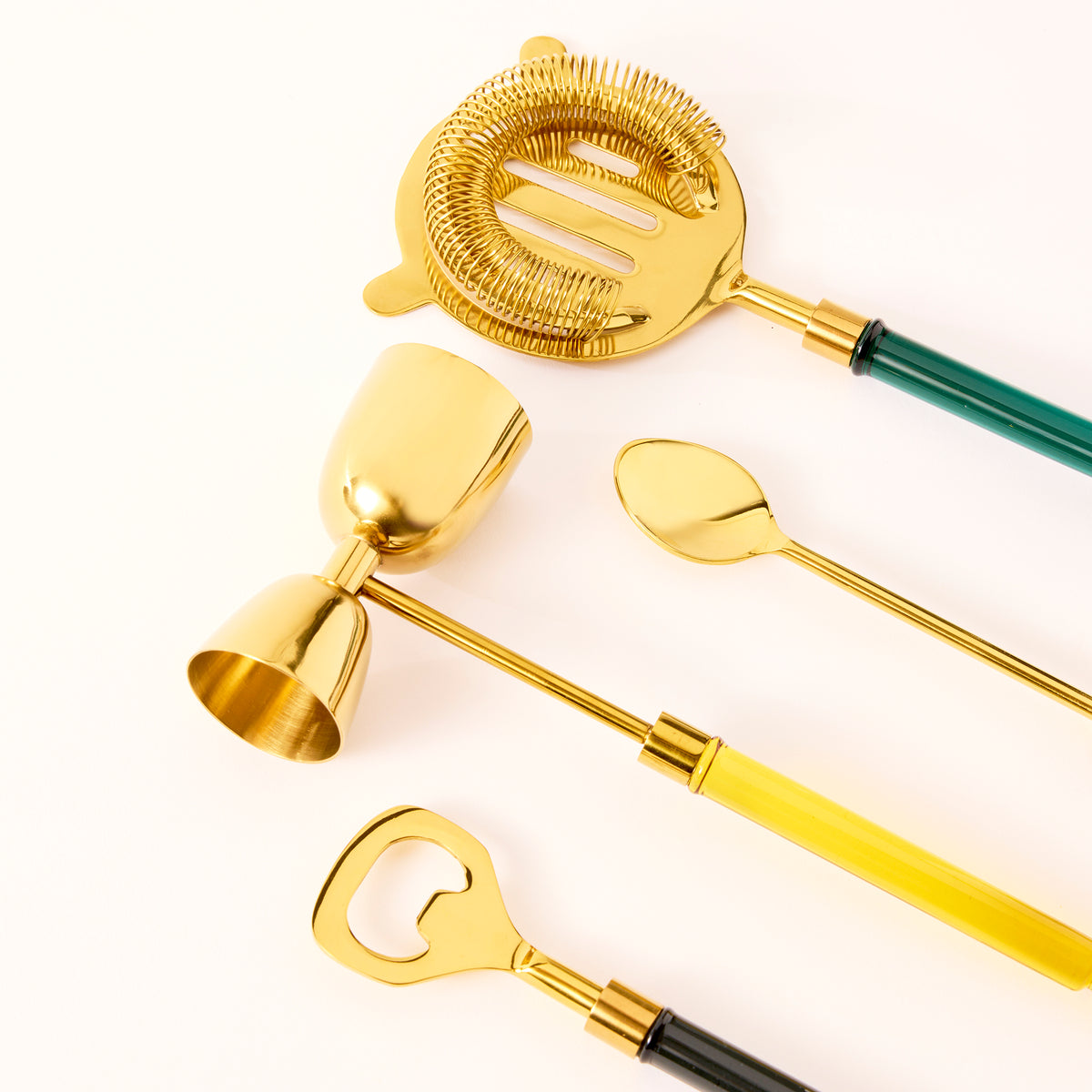 Set of gold cocktail tools including a strainer, jigger, and bottle opener on a white background
