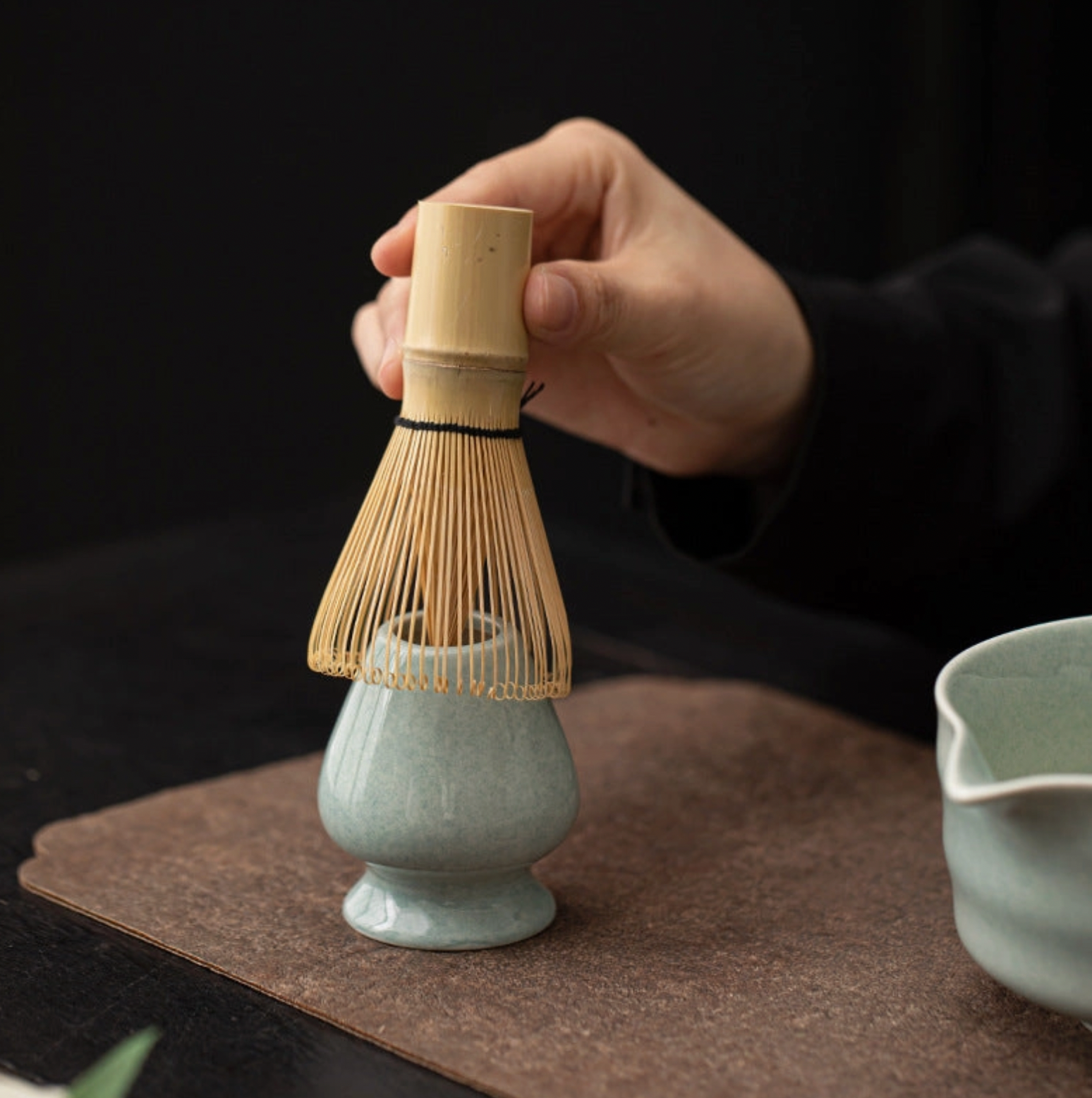 Person holding a wooden whisk over a ceramic container on a dark surface