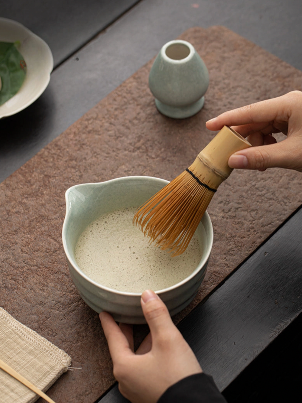 Person preparing matcha in a ceramic bowl with a whisk on a wooden surface.