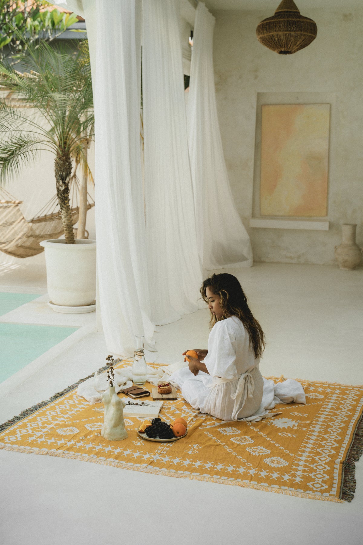 Woman sitting on a yellow rug by a poolside with white curtains and a palm tree in the background.
