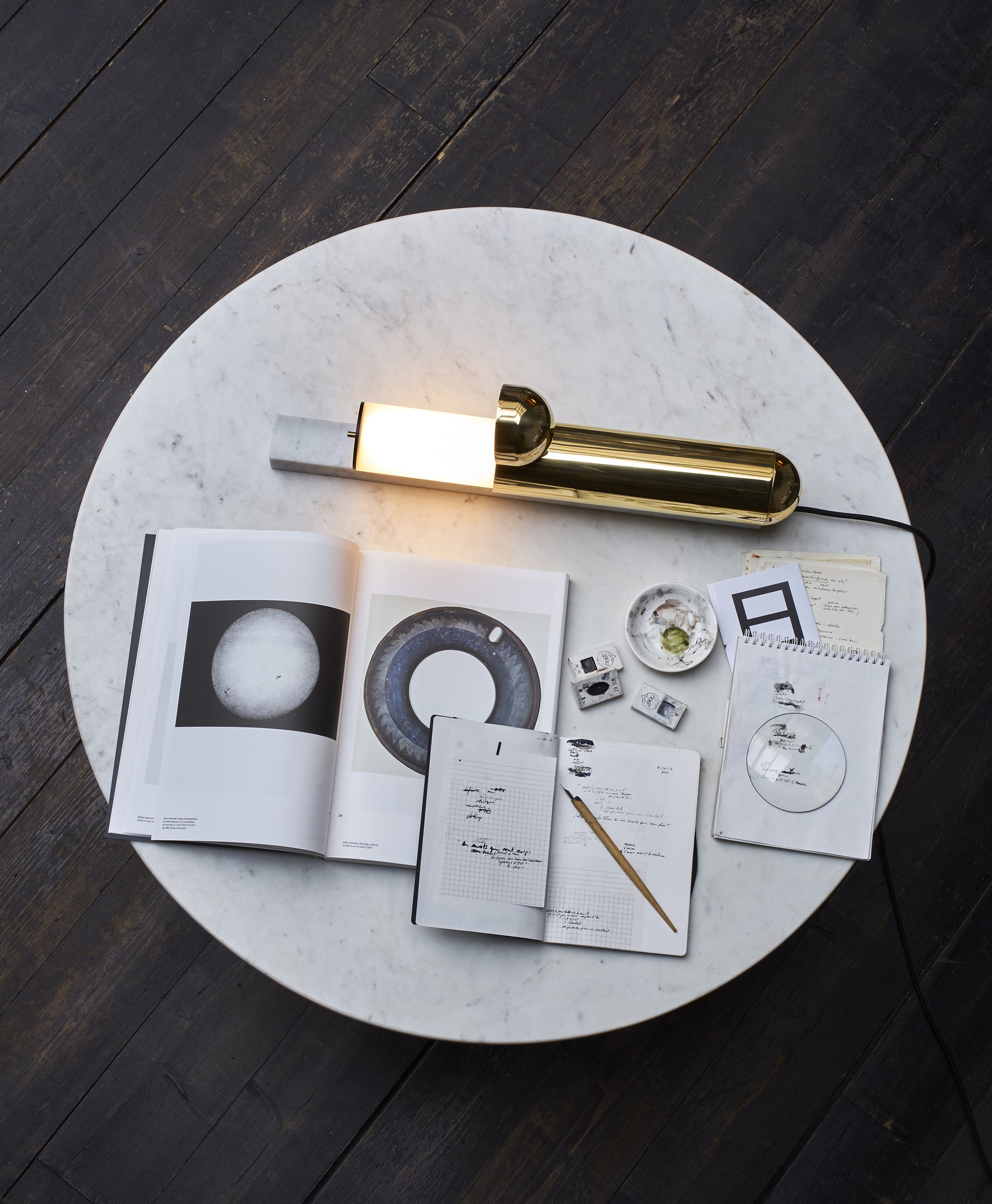 Round marble table with decorative items including a gold lamp and printed materials on a dark wooden floor.