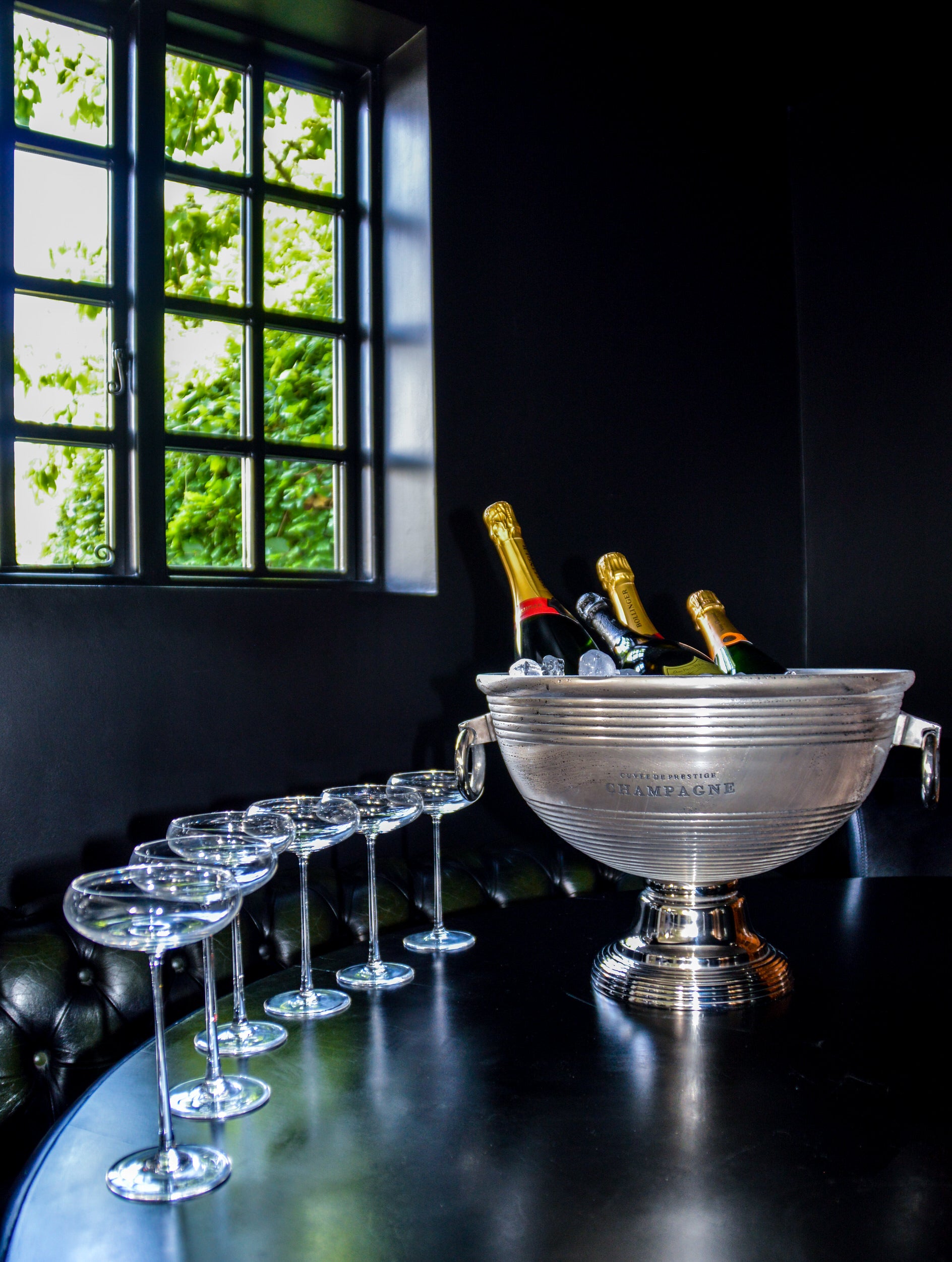 Silver champagne bucket with bottles and glasses on a dark surface near a window.