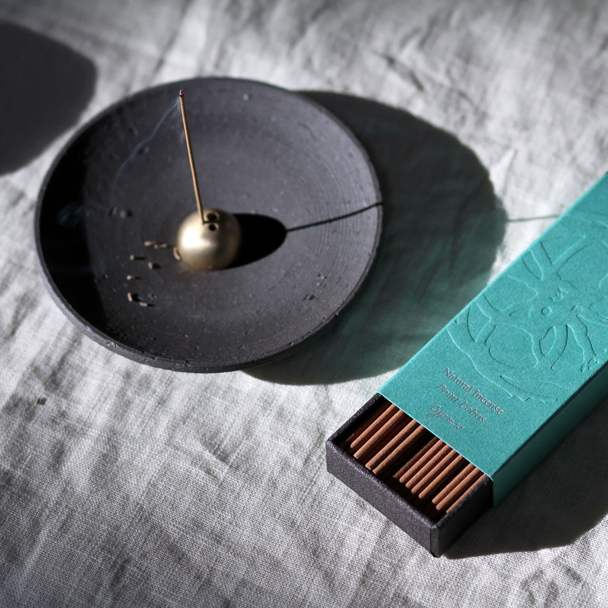Incense holder with a gold incense stick and a teal box of incense on a textured surface.