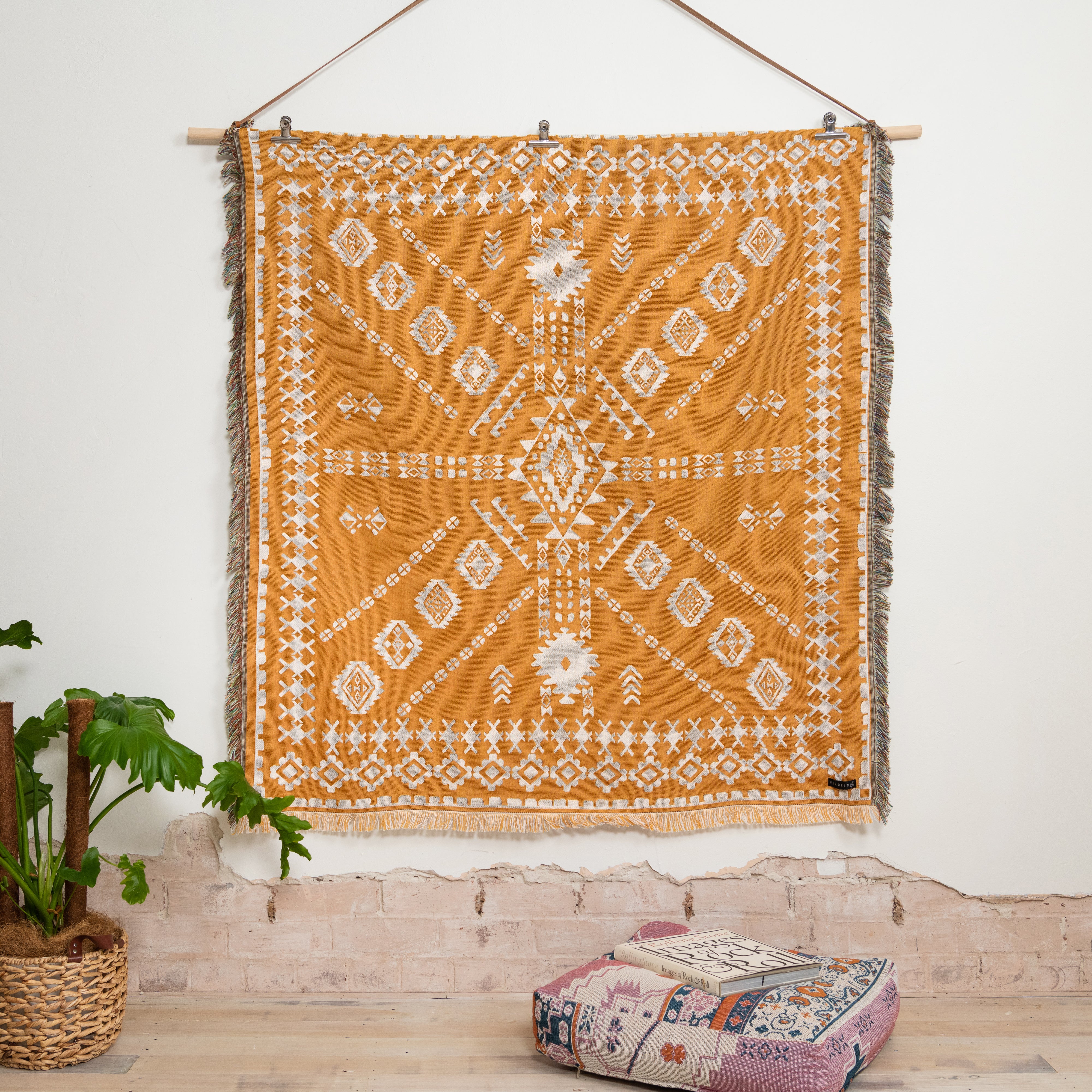 Decorative tapestry with geometric pattern hanging on a white wall, with a plant and cushion in the foreground.