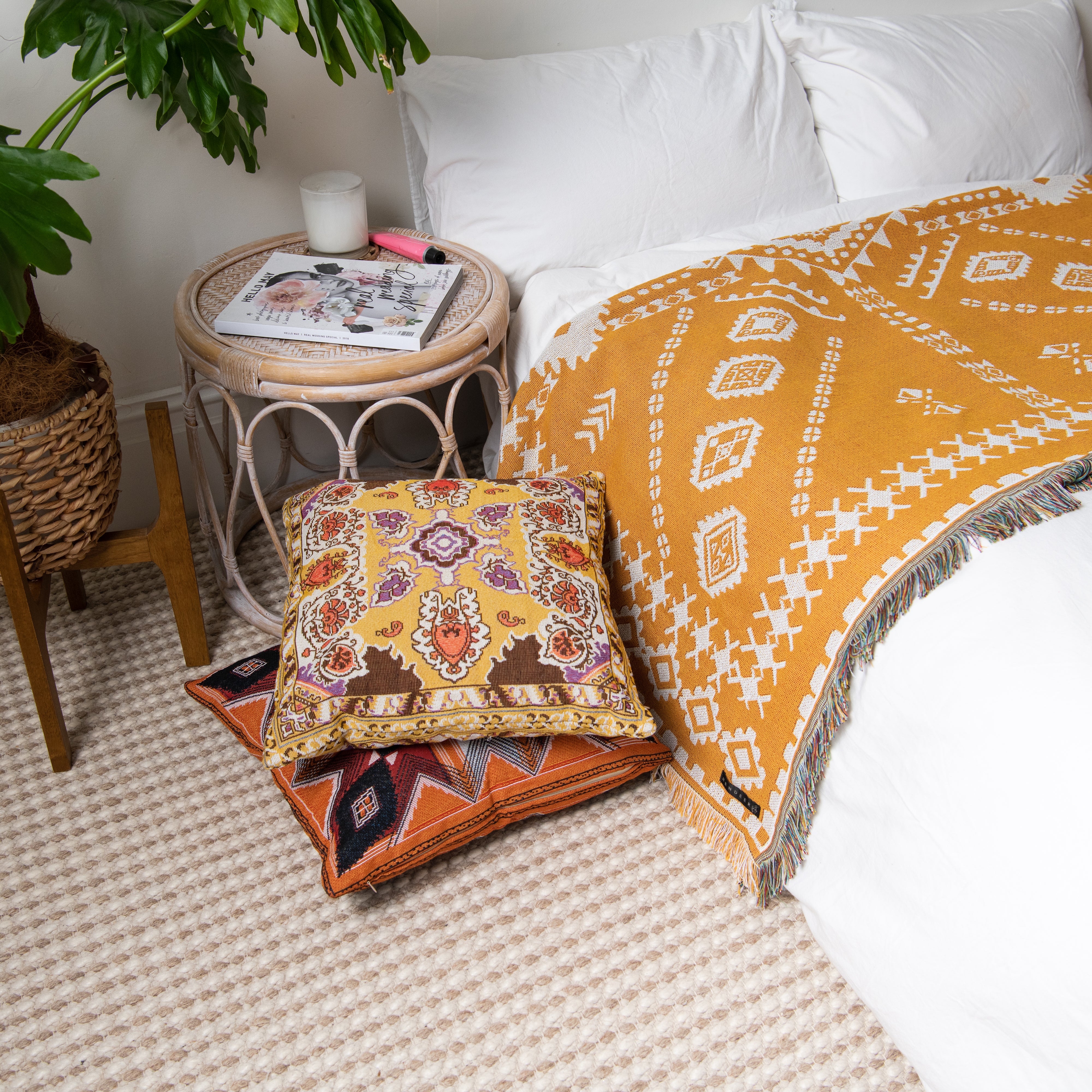 Decorative pillows and a blanket on a bed with a side table and plant in the background.