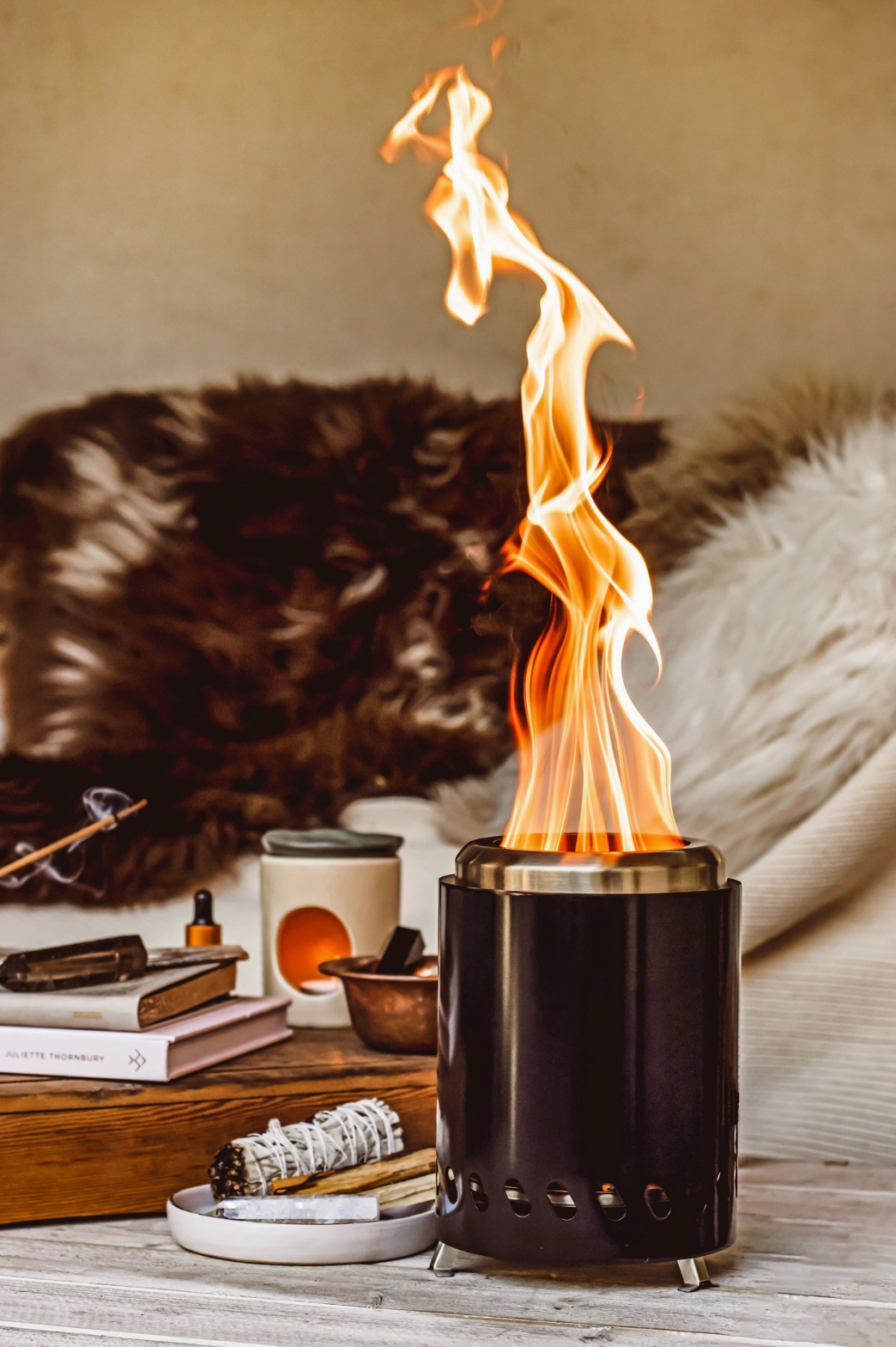 Black metal fire pit with flames on a wooden surface with books and candles in the background.