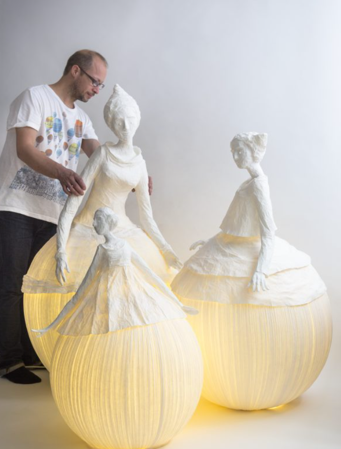 Man interacting with large paper mache sculptures of women in dresses on a white background
