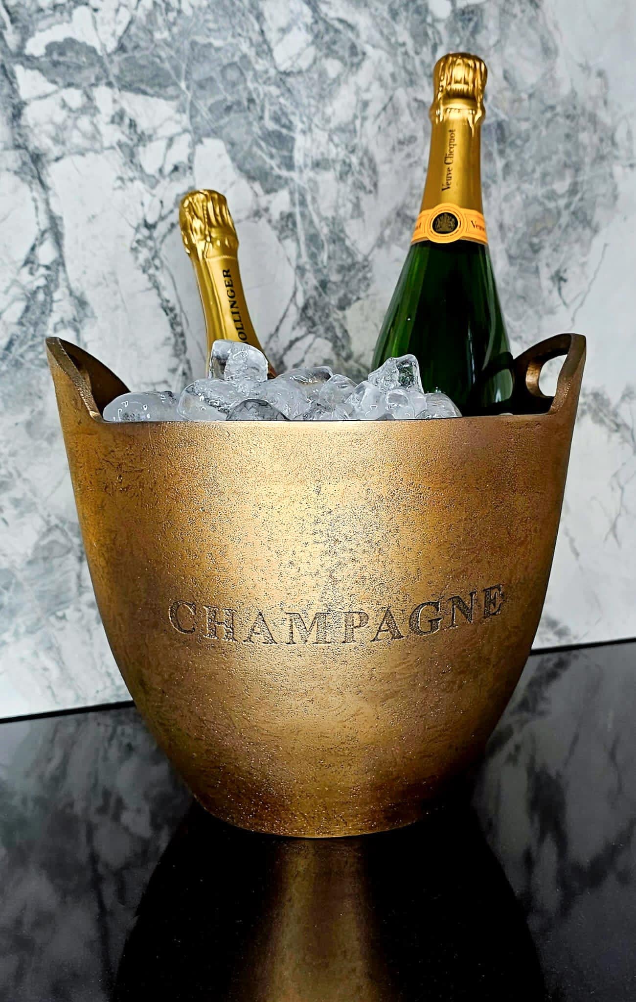 Two champagne bottles in a bronze 'Champagne' ice bucket against a marble background