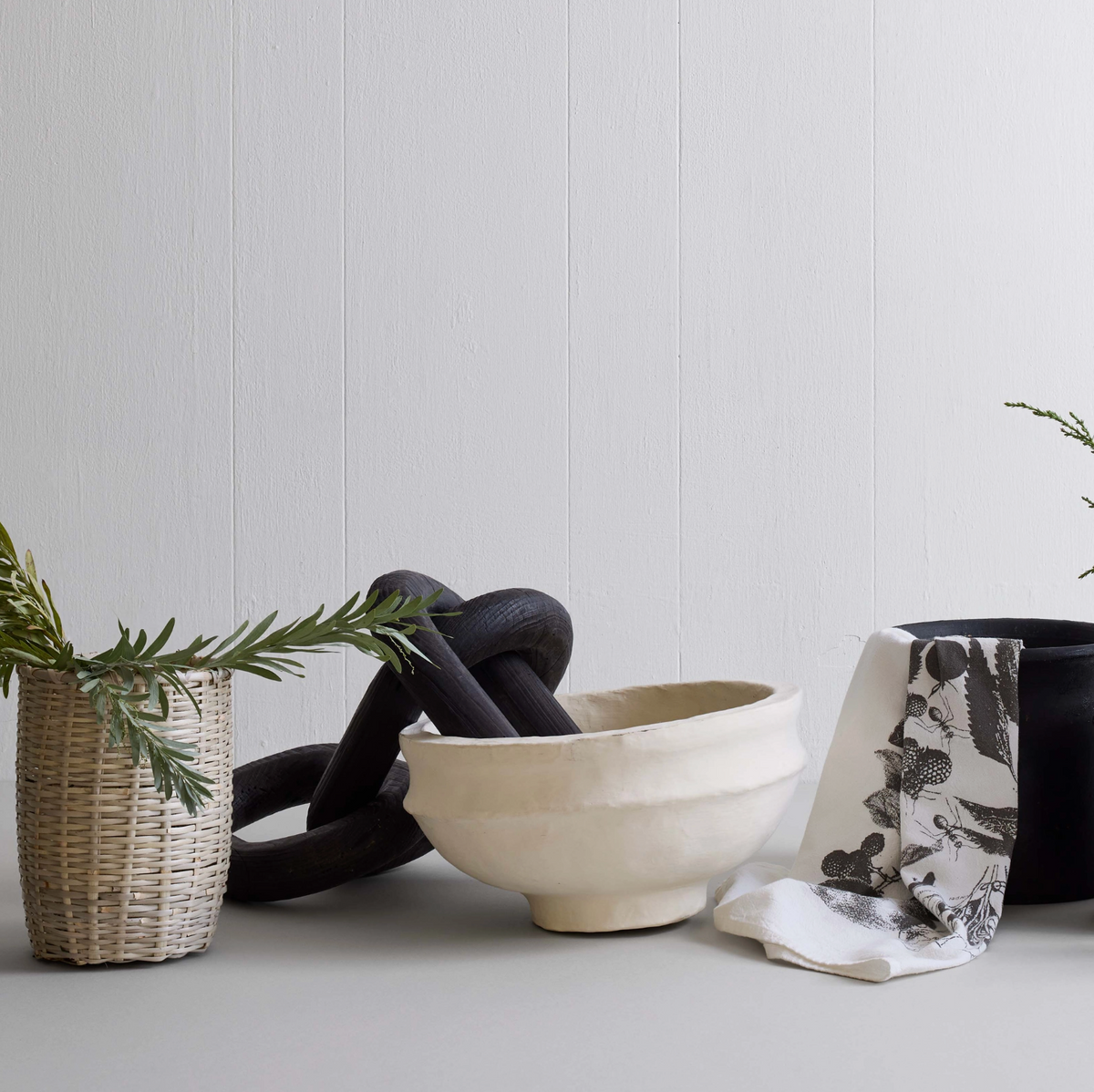 Decorative items including a woven basket, black sculpture, ceramic bowl, and patterned towel on a light surface with a white paneled wall background.