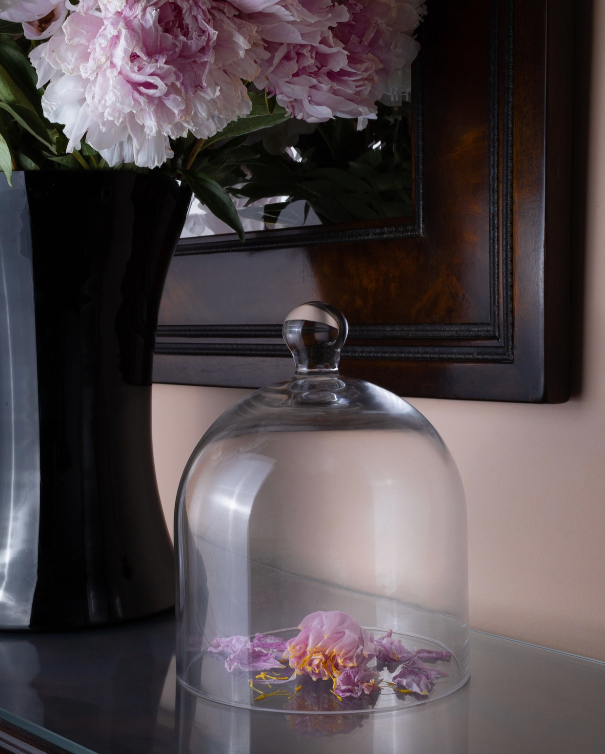 Glass cloche with flowers on a surface, with a vase of pink flowers in the background.