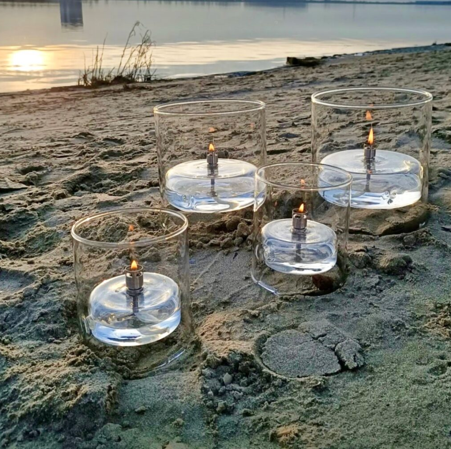 Glass candle holders with candles on a sandy beach at sunset.
