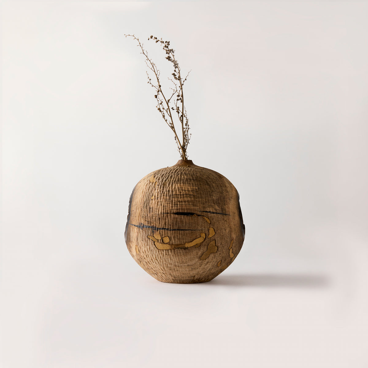Wooden vase with a textured surface and dried plant on a light gray background