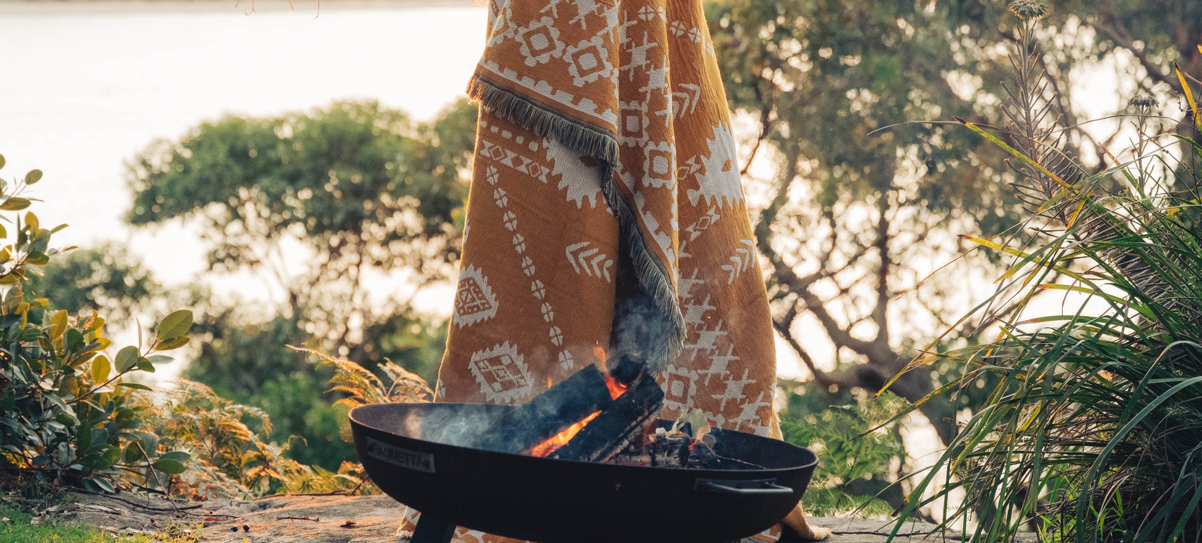 Person wrapped in a patterned blanket by a campfire with a scenic background