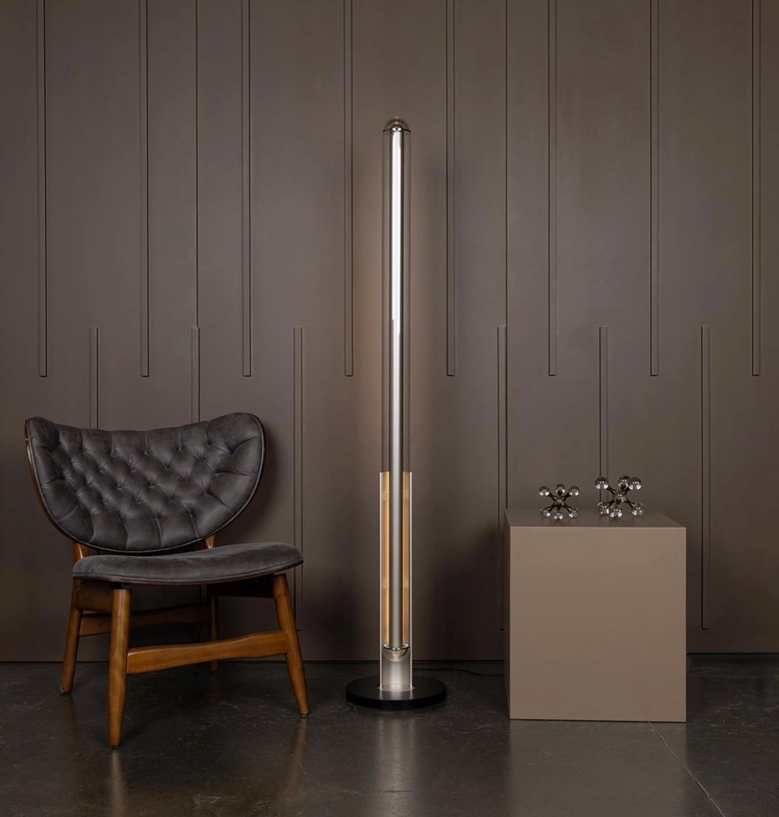 Tall cylindrical floor lamp next to a dark gray chair against a brown paneled wall.