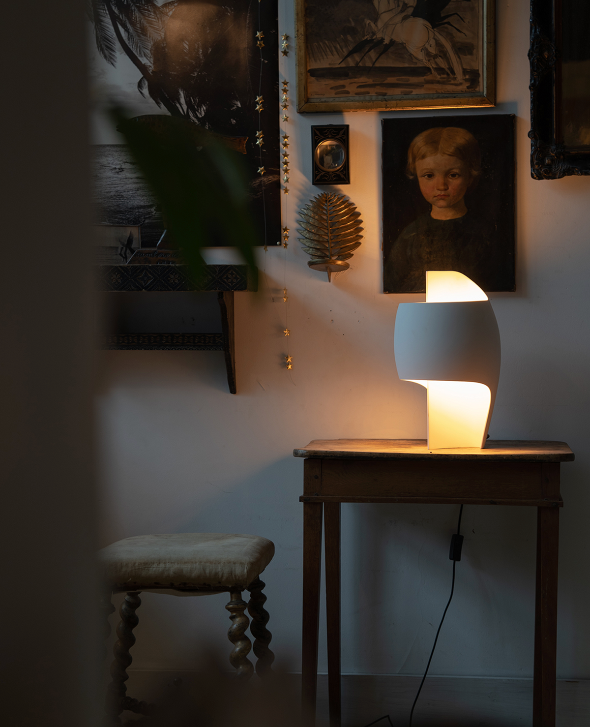 Lamp on a wooden table with framed pictures on the wall in a dimly lit room.