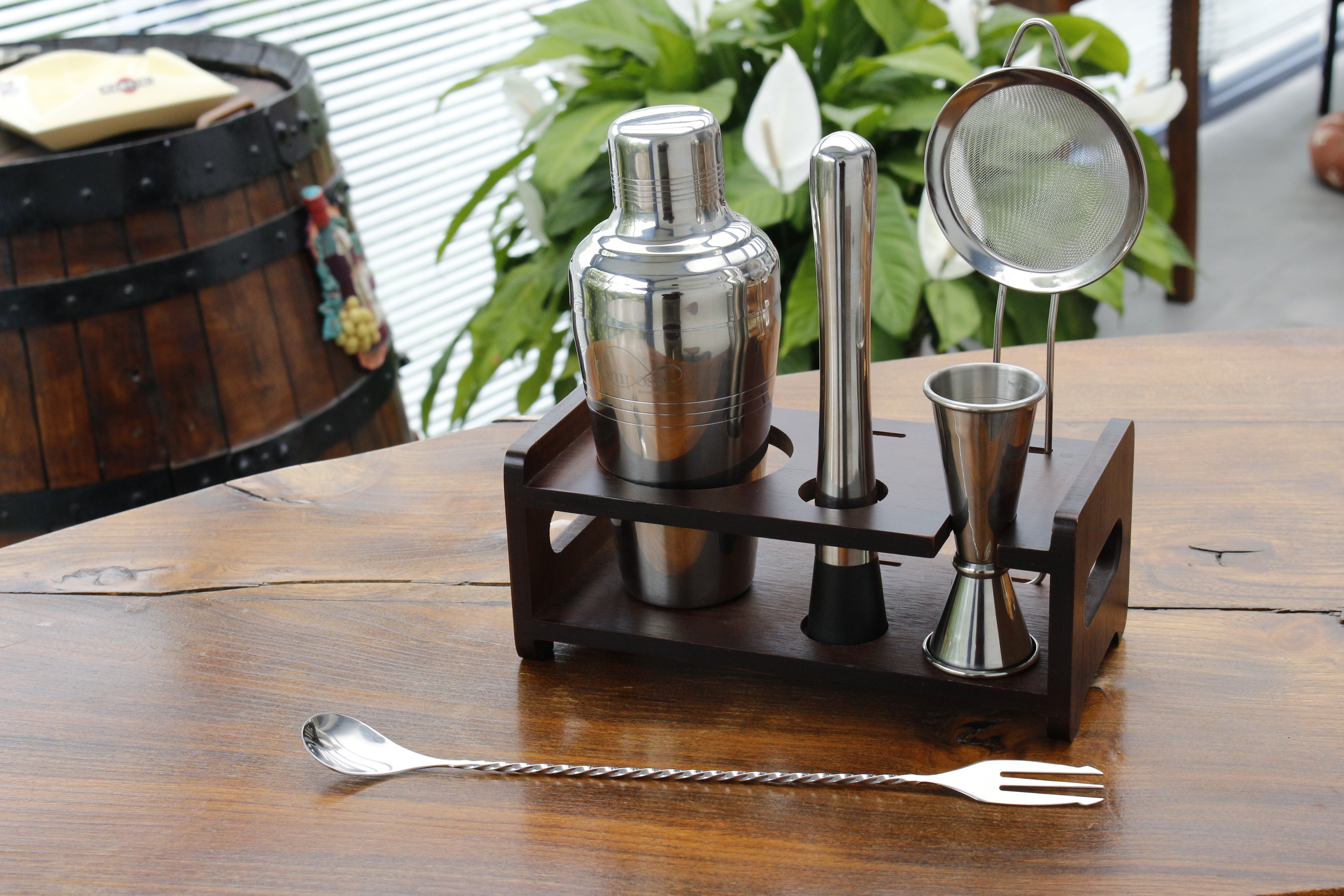 Set of cocktail-making tools including a shaker, jigger, and strainer on a wooden stand with a blurred background.