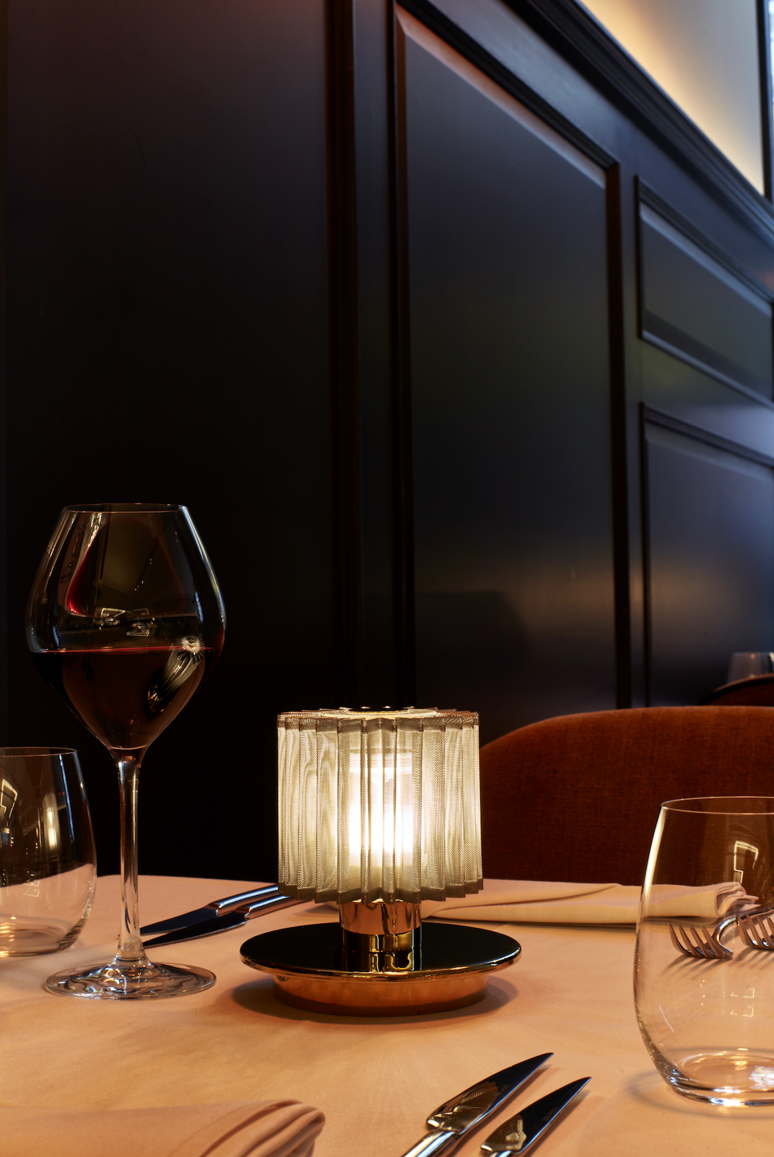 Dining table setting with a small lamp, wine glasses, and cutlery against a dark wall.