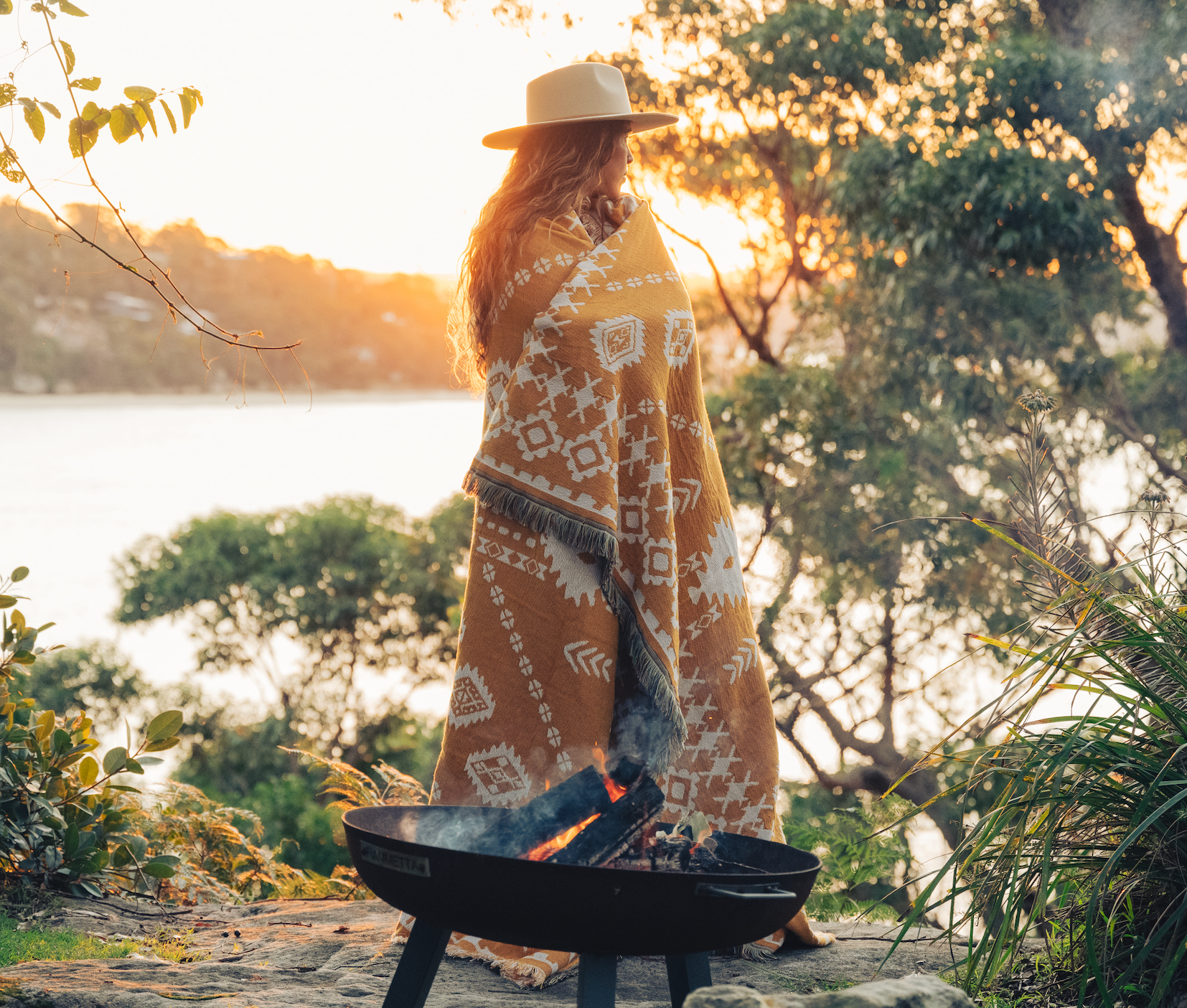 Person wrapped in a patterned blanket standing by a fire pit with a scenic background