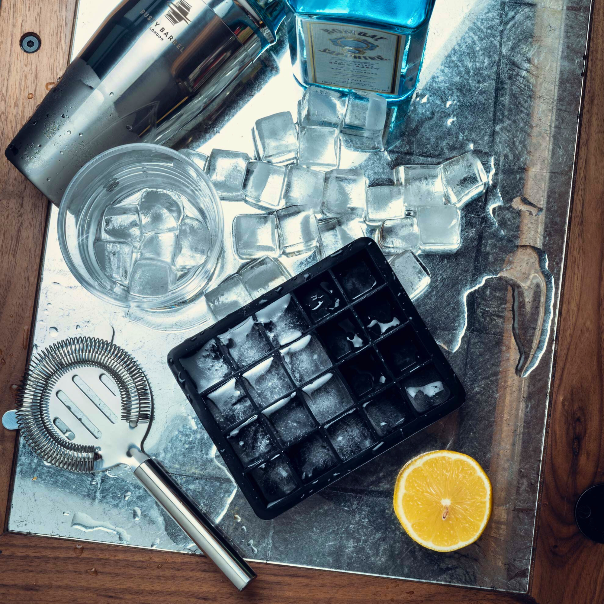 Bartending setup with ice cubes, bottle, glass, and lemon on a metallic surface.