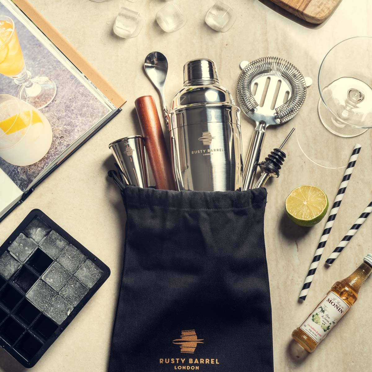 Cocktail-making tools and ingredients on a marble surface with a 'Rusty Barrel London' pouch.