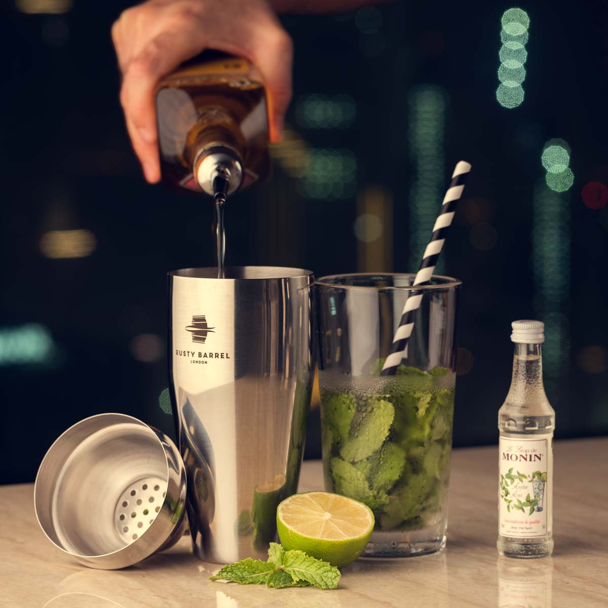 Person pouring a drink into a cocktail shaker with a glass of ice and mint leaves on a bar counter.