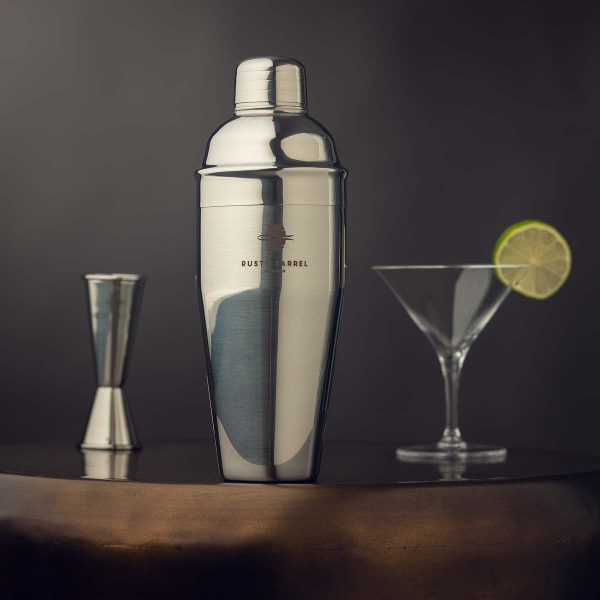 Silver cocktail shaker with a glass, jigger, and lime wedge on a dark surface.