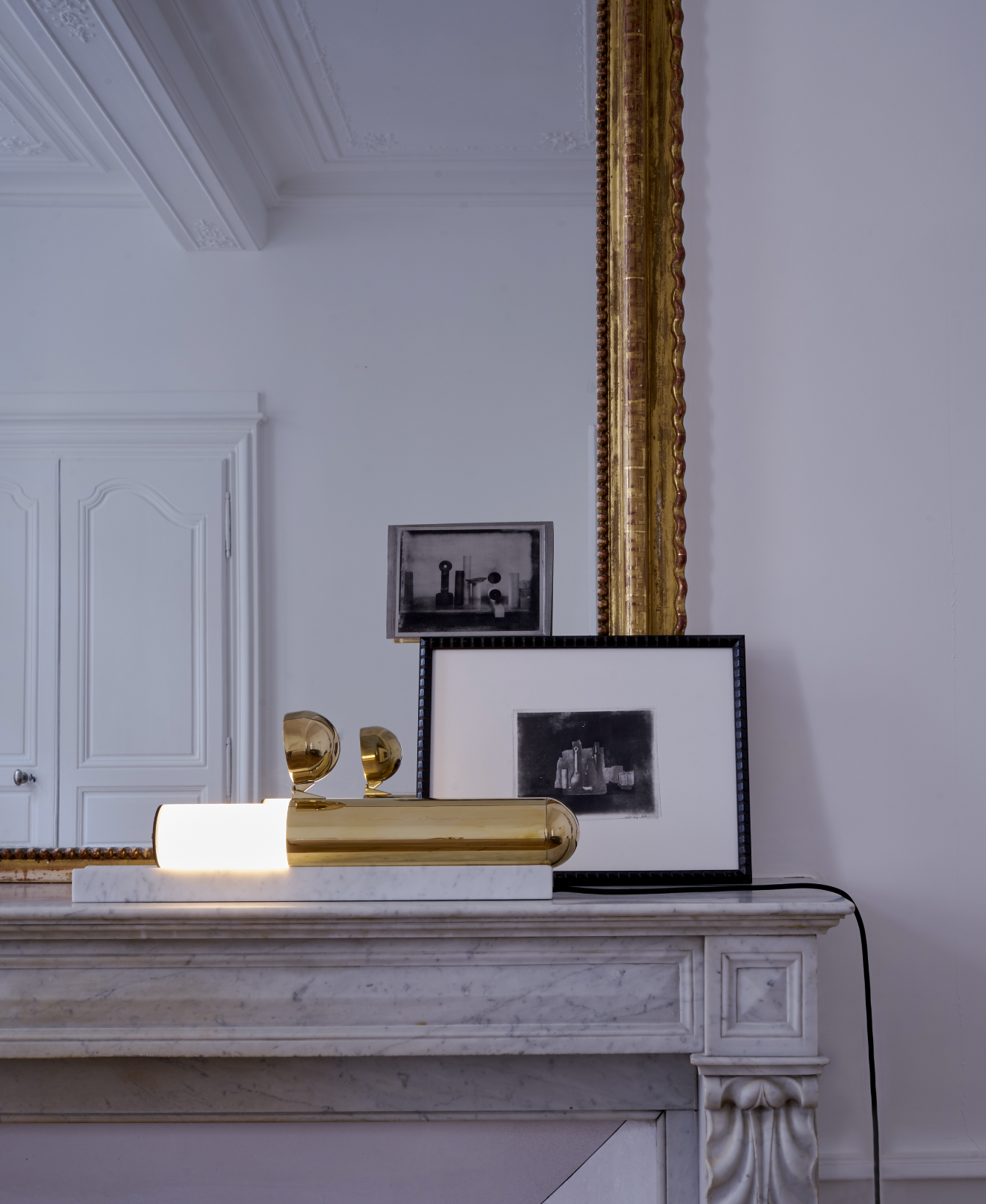 Gold lamp and framed pictures on a white mantelpiece with a large mirror in the background.