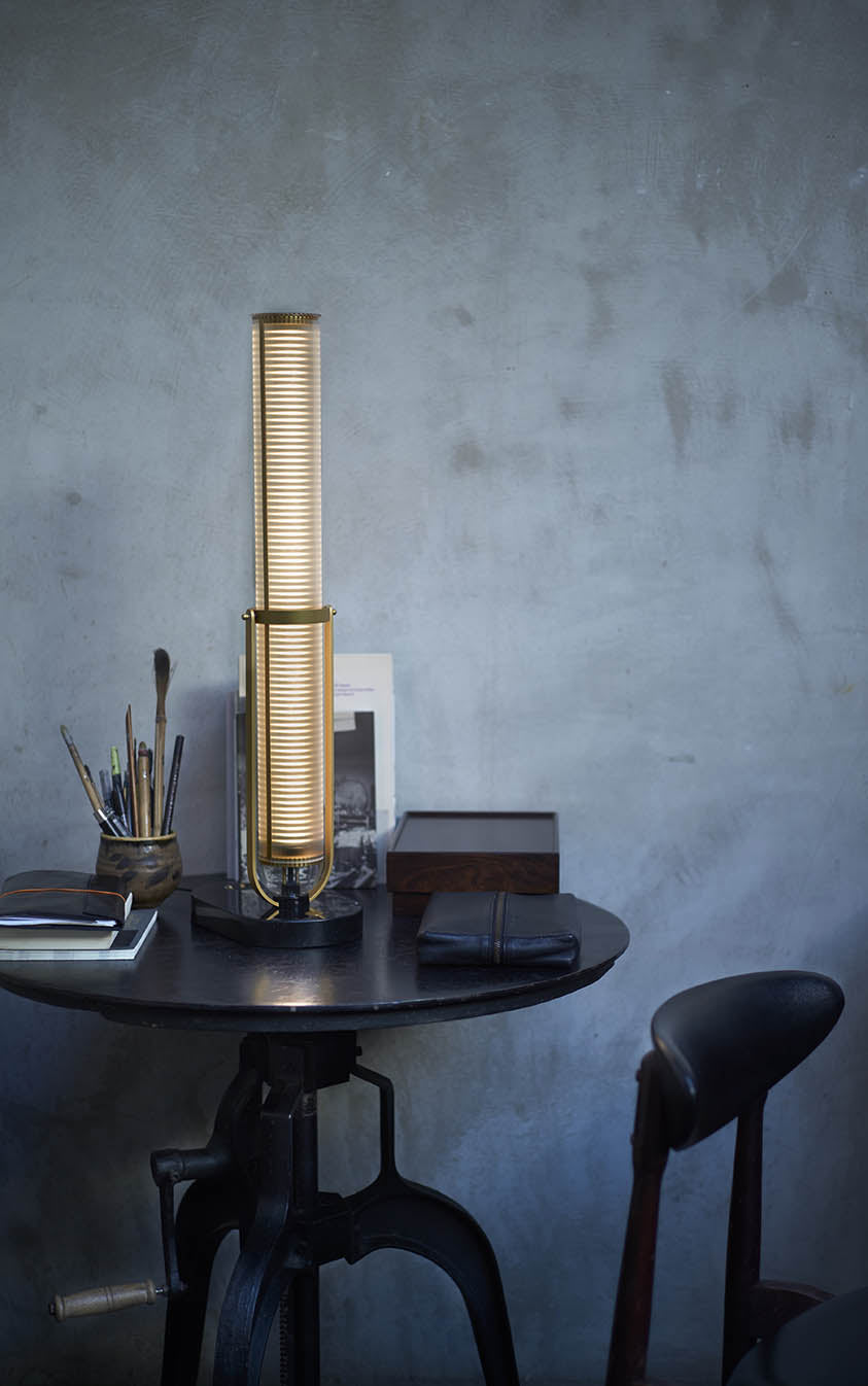 Tall cylindrical lamp on a dark wooden table against a textured gray wall.