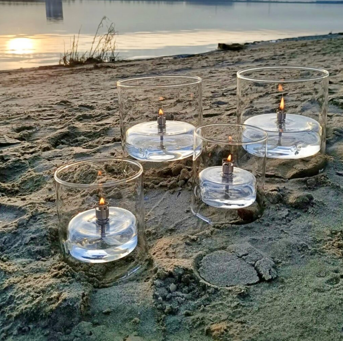 Glass candle holders with candles on a sandy beach at sunset.