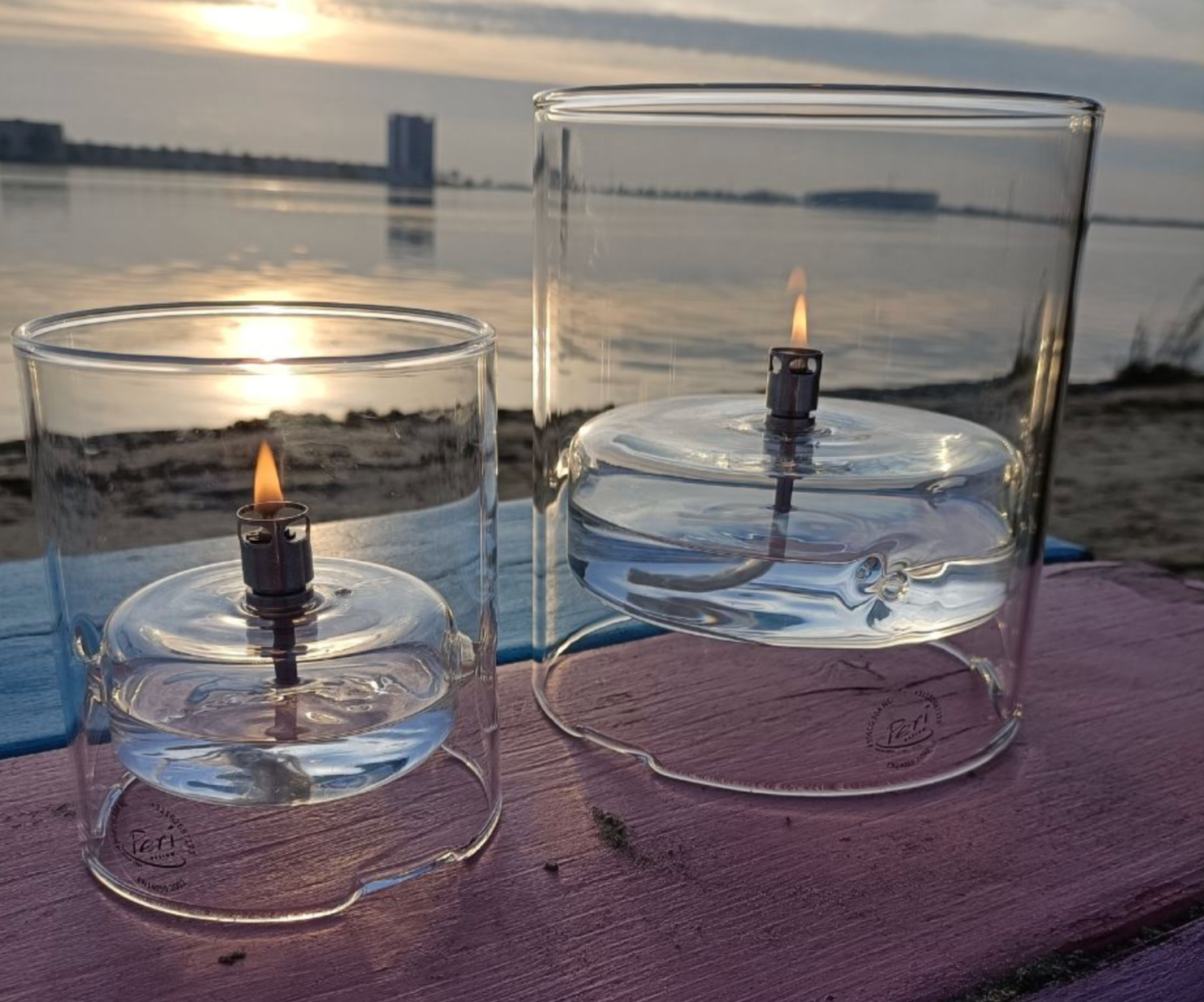 Two glass lanterns with candles on a wooden surface by a waterfront at sunset.