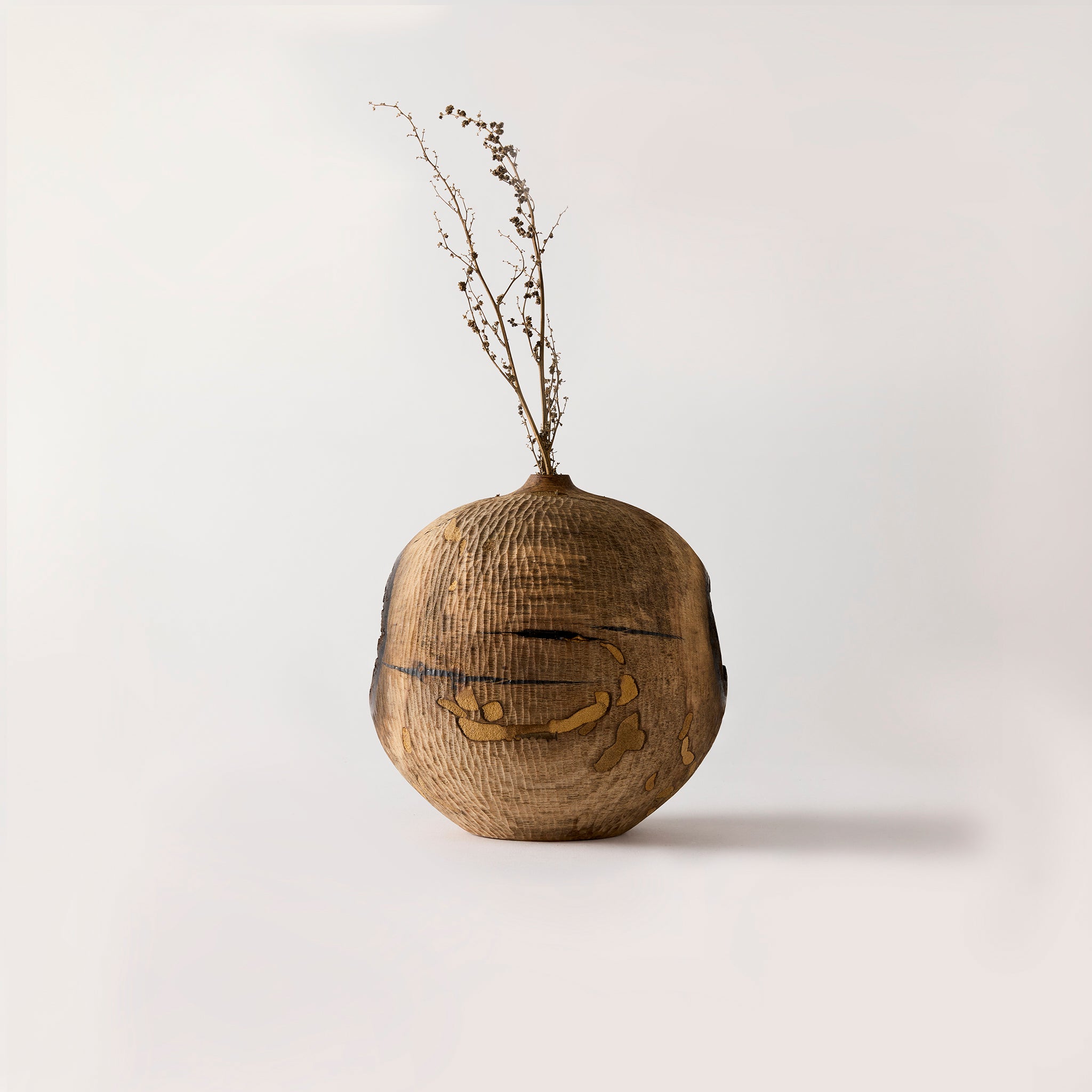 Wooden vase with a textured surface and dried plant on a light gray background